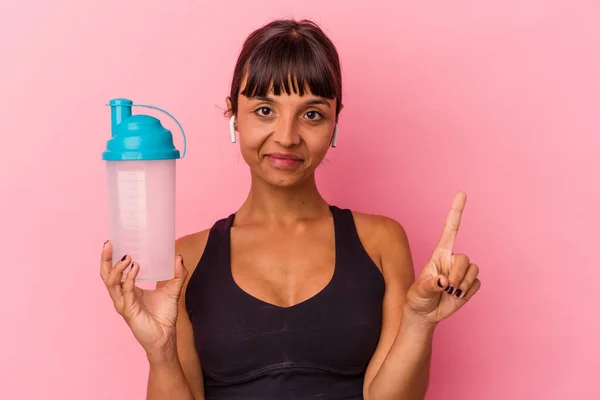 ピンクの背景に単離されたタンパク質シェイクを飲む若い混合人種の女性は指でナンバーワンを示す — ストック写真