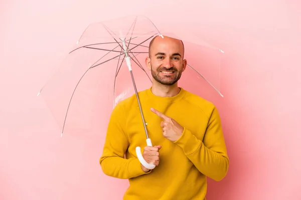 ピンクの背景に孤立した傘を持っている若い白人のはげ男は笑顔と脇を指して 空白のスペースで何かを示す — ストック写真
