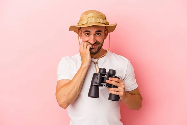 年轻的高加索秃头男子拿着双筒望远镜 用粉红的背景隔离 咬着指甲 紧张而焦虑 — 图库照片