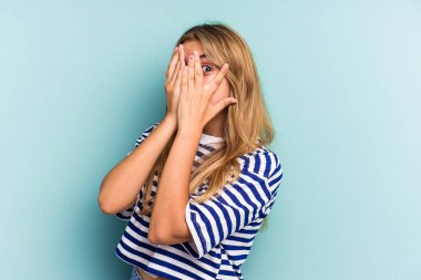Mavi arka planda izole edilmiş beyaz tenli genç bir kadın parmaklarıyla gözünü kırp korkmuş ve gergin..