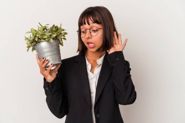 Beyaz arka planda bir bitki tutan genç melez iş kadını bir dedikoduyu dinlemeye çalışıyor..