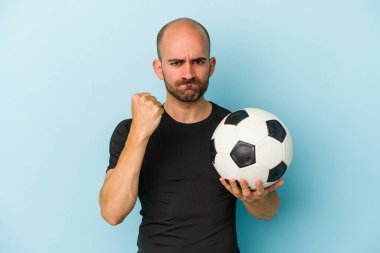 Genç iş adamı kel adam mavi arka planda izole edilmiş bir şekilde futbol oynuyor. Kameraya yumruk gösteriyor, agresif yüz ifadesi var..