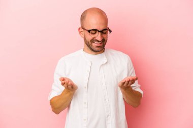 Beyaz, kel bir adam pembe arka planda izole edilmiş. Elinde avuç içi olan bir şeyle kameraya sunuyor..