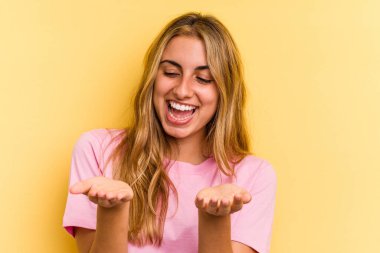 Sarışın, sarı arka planda izole edilmiş genç bir kadın elinde avuç içi bir şeyle kameraya sunuyor..