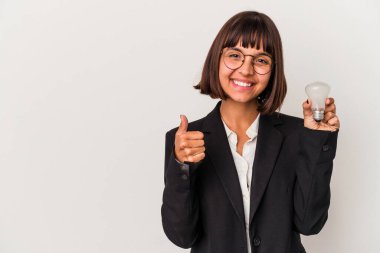 Beyaz arka planda izole edilmiş ampulü tutan genç, melez bir iş kadını gülümsüyor ve başparmağını kaldırıyor.