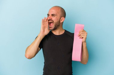 Genç sporcu kel adam mavi arka planda izole edilmiş elastik bir bant tutuyor bağırıyor ve avuç içini açık tutuyor..