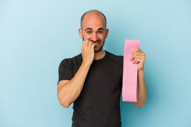 Genç sporcu kel bir adam mavi arka planda izole edilmiş elastik bir bant tutuyor tırnaklarını yiyor, gergin ve çok endişeli..