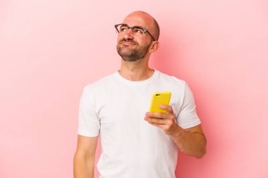 Kafkasyalı kel adam cep telefonunu pembe arka planda izole bir şekilde tutarak hedeflerine ve amaçlarına ulaşmayı hayal ediyor.