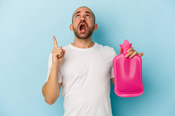 Молодой лысый мужчина держит горячий мешок с водой на синем фоне, указывая вверх ногами с открытым ртом.