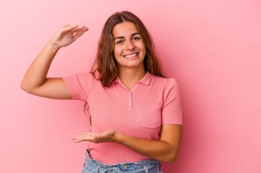 Pembe arka planda izole edilmiş genç beyaz bir kadın işaret parmaklarıyla küçük bir şey tutuyor, gülümsüyor ve kendinden emin..