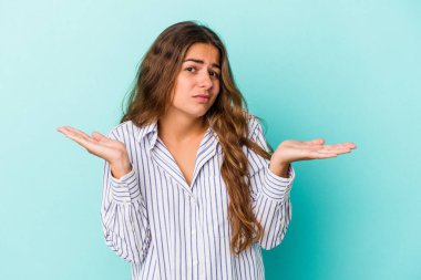 Kafkasyalı genç bir kadın mavi arka planda izole edilmiş sorgulama sırasında omuz silkiyor ve kuşkulanıyor..