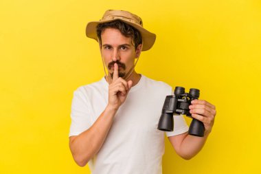 Sarı arka planda dürbün tutan genç beyaz adam bir sır saklıyor ya da sessizlik istiyor..