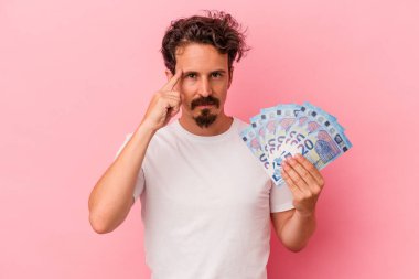 Elinde banknotlar tutan beyaz bir adam pembe arka planda izole edilmiş, parmağıyla tapınağı işaret eden, düşünen, bir göreve odaklanmış..