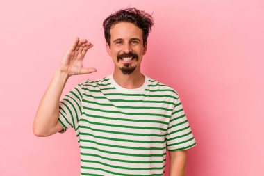 Pembe arka planda izole edilmiş genç beyaz adam işaret parmaklarıyla küçük bir şey tutuyor, gülümsüyor ve kendinden emin..