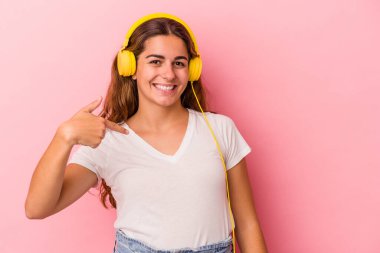 Pembe arka planda izole edilmiş müzik dinleyen beyaz bir kadın gömlek fotokopi alanını elle işaret ediyor, gururlu ve kendinden emin.