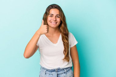 Mavi arka planda izole edilmiş genç beyaz kadın gülümsüyor ve başparmağını kaldırıyor.