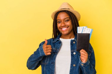 Sarı arka planda izole edilmiş pasaportu olan genç Afrikalı Amerikalı kadın gülümsüyor ve başparmağını kaldırıyor.