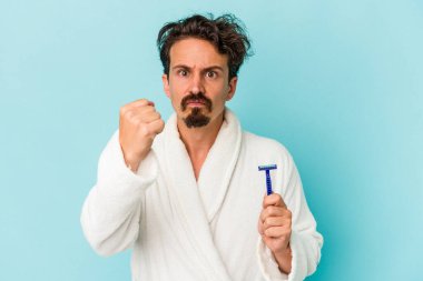 Mavi arka planda jilet tutan genç beyaz bir adam kameraya yumruk gösteriyor, agresif yüz ifadesi..