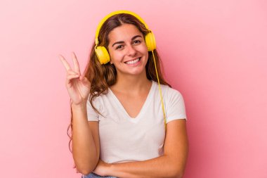 Pembe arka planda müzik dinleyen beyaz bir kadın iki numarayı parmaklarıyla gösteriyor..