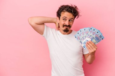 Elinde banknotlar tutan beyaz bir adam pembe arka planda izole edilmiş kafasının arkasına dokunuyor, düşünüyor ve bir seçim yapıyor..