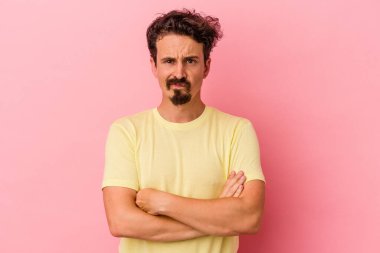 Pembe arka planda izole edilmiş genç beyaz adam hoşnutsuzca kaşlarını çatmış, kollarını kavuşturmuş..