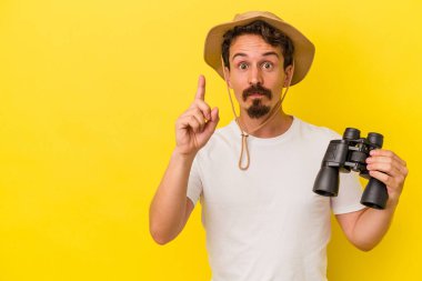 Dürbün tutan genç beyaz adam sarı arka planda bir numarayı parmağıyla gösteriyor..