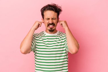 Pembe arka planda izole edilmiş genç beyaz bir adam parmaklarıyla kulaklarını kapatıyor, gürültülü bir ortam yüzünden stresli ve çaresiz..
