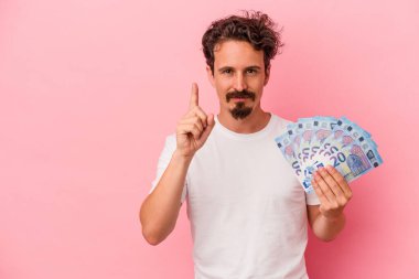 Elinde banknotlar tutan beyaz bir adam pembe arka planda bir numarayı parmağıyla gösteriyor..