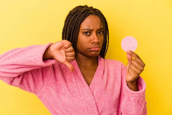 年轻的非洲裔美国女人穿着浴衣 手里拿着一个用黄色背景隔开的化妆盘 表现出不喜欢的姿态 大姆指朝下 不同意的概念 — 图库照片