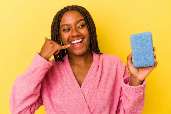 身穿浴衣的年轻非洲裔美国女人拿着用黄色背景隔开的蓝色海绵 用手指打了一个手机手势 — 图库照片