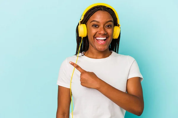 青い背景の上で孤立した音楽を聴いている若いアフリカ系アメリカ人の女性が笑いながら指摘し 何もない空間で何かを示す — ストック写真