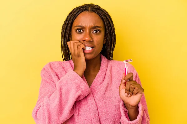 身穿浴衣的年轻非洲裔美国女人拿着一把用黄色背景隔离的剃须刀刀片咬着指甲 既紧张又焦虑 — 图库照片