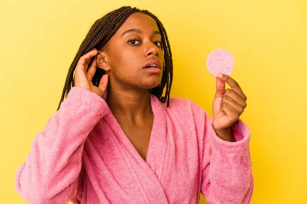 Young african american woman wearing a bathrobe holding a make up disk isolated on yellow background  trying to listening a gossip.