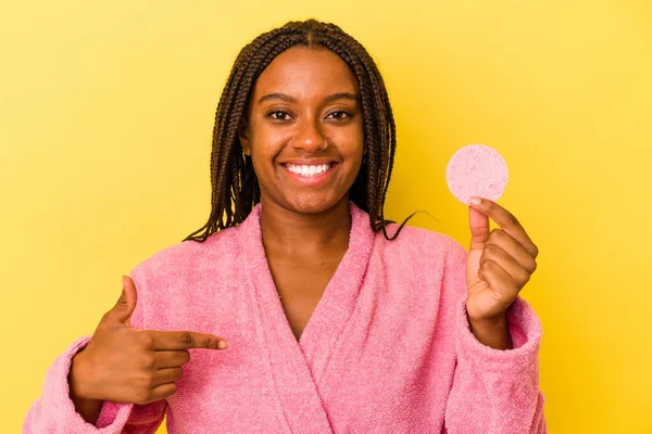 身穿浴衣的年轻的非洲裔美国女人拿着一个用黄色背景的人隔开的化妆品盘 手拉手指着衬衫的复制品空间 自豪而自信 — 图库照片
