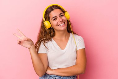Pembe arka planda izole edilmiş müzik dinleyen beyaz bir kadın neşeli ve kaygısız parmaklarıyla bir barış sembolü gösteriyor..