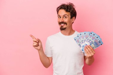 Elinde banknotlar tutan beyaz bir adam pembe arka planda izole edilmiş gülümseyen ve işaret eden boş alanda bir şey gösteriyor..
