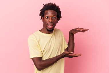 Genç Afrikalı Amerikalı bir adam pembe arka planda izole edilmiş. Elleri arasında bir kopya alanı tutarken şok ve hayret içinde..