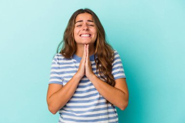 Mavi arka planda izole edilmiş genç beyaz bir kadın el ele tutuşup ağza yakın dua ediyor, kendinden emin..