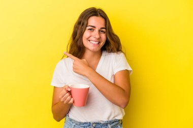 Sarı arka planda izole edilmiş bir kupa tutan genç beyaz kadın gülümsüyor ve boş alanda bir şeyler gösteriyor..
