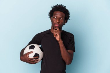 Amerikan futbolu oynayan genç Afro-Amerikalı adam mavi arka planda izole edilmiş şüpheli ve şüpheci bir ifadeyle yana bakıyor..