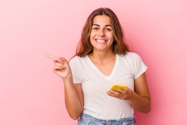 Pembe arka planda izole edilmiş cep telefonu kullanan beyaz bir kadın gülümsüyor ve boş alanda bir şeyler gösteriyor..