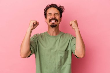 Pembe arka planda izole edilmiş genç beyaz bir adam zaferi, tutkuyu ve coşkuyu kutluyor, mutlu bir ifade..