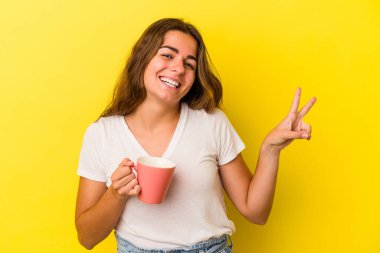 Kafkasyalı genç bir kadın sarı arka planda izole edilmiş bir kupa tutuyor neşeli ve tasasız parmaklarıyla barış sembolü gösteriyor..
