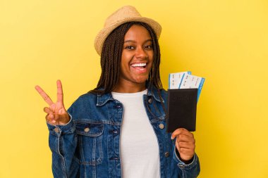 Pasaportu olan genç Afrikalı Amerikalı kadın sarı arka planda izole edilmiş neşeli ve kaygısız parmakları olan bir barış sembolü gösteriyor..