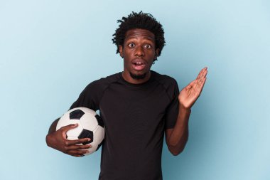 Futbol oynayan genç Afro-Amerikalı adam mavi arka planda izole edilmiş, şaşırmış ve şok olmuş..