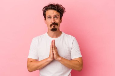 Pembe arka planda izole edilmiş genç beyaz bir adam. El ele tutuşup, ağız kenarında dua ediyor. Kendine güveniyor..