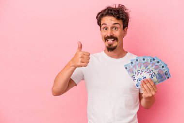 Pembe arka planda izole edilmiş banknotları tutan genç beyaz adam gülümsüyor ve başparmağını kaldırıyor.