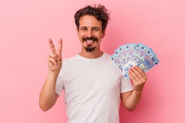 Elinde banknotlar tutan beyaz bir adam pembe arka planda iki numarayı parmaklarıyla gösteriyor..