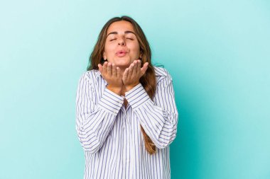 Mavi arka planda izole edilmiş genç, beyaz bir kadın. Dudaklarını kıvırıyor ve avuç içlerine hava öpücüğü yolluyor..