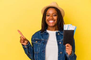 Sarı arka planda izole edilmiş pasaportu olan genç Afrikalı Amerikalı kadın gülümsüyor ve boş alanda bir şey gösteriyor..